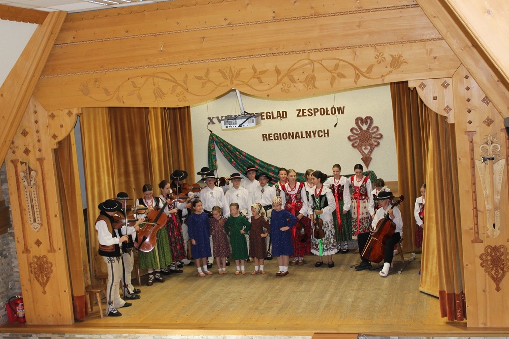 Drewniana scena na której stoją ubrani w góralskie stroje muzycy i tancerze