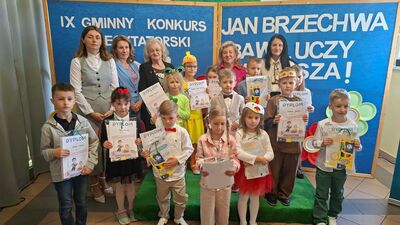 Zdjęcie grupowe na tle konkursowej dekoracji. W przedniej części stoi grupa kilkanaściorga dzieci w różnym wieku, trzymających pamiątkowe dyplomy oraz nagrody rzeczowe. Niektóre dzieci są w strojach galowych, inne mają elementy kostiumów (np. maskę lwa, opaskę z ptakiem). W tylnym rzędzie za dziećmi stoi pięć dorosłych kobiet – członkiń jury.