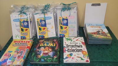 Zdjęcie przedstawia stół przykryty zielonym obrusem, na którym ułożone są nagrody dla uczestników konkursu. Widoczne są pakiety zapakowane w przezroczystą folię i przewiązane zieloną wstążką, zawierające kolorowanki oraz zestawy kredek ołówkowych marki Astra. Obok leżą ułożone w stosy książki z bajkami i wierszami, w tym m.in. „Jan Brzechwa dzieciom” oraz „Julian Tuwim – Wiersze”.