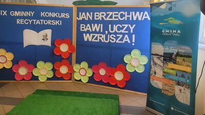Zdjęcie przedstawia pustą scenografię przygotowaną na wydarzenie. Głównym elementem jest niebieska, dwuskrzydłowa ścianka z białymi napisami „IX GMINNY KONKURS RECYTATORSKI” oraz „JAN BRZECHWA BAWI, UCZY, WZRUSZA!”. Dekorację zdobią duże, kolorowe kwiaty wykonane z papierowych talerzyków oraz element w kształcie otwartej książki. Przed ścianką leży zielony podest imitujący trawę, a po prawej stronie stoi baner roll-up z logo Gminy Nowy Targ.