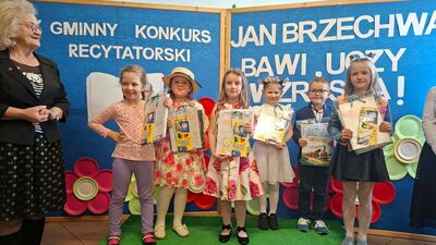 Zdjęcie przedstawia sześcioro dzieci (pięć dziewczynek i jednego chłopca) stojących na zielonym podwyższeniu podczas wręczenia nagród. Pozują na tle niebieskiej ścianki z hasłem konkursu o Janie Brzechwie. Dzieci trzymają w rękach pakiety z nagrodami (m.in. kredki Astra) oraz książki. Po lewej stronie stoi starsza kobieta w czarnej spódnicy w grochy, patrząca z uśmiechem na uczestników.