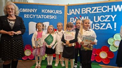 Grupa pięciu dziewczynek stoi na podwyższeniu pokrytym sztuczną trawą na tle niebieskiej dekoracji z napisem „GMINNY KONKURS RECYTATORSKI JAN BRZECHWA BAWI, UCZY, WZRUSZA!”. Dzieci trzymają w rękach nagrody w postaci książek i dyplomów; jedna z dziewczynek ubrana jest w biały fartuch lekarski ze stetoskopem, inna ma na głowie opaskę z czułkami biedronki. Po lewej stronie stoi uśmiechnięta członkini jury.